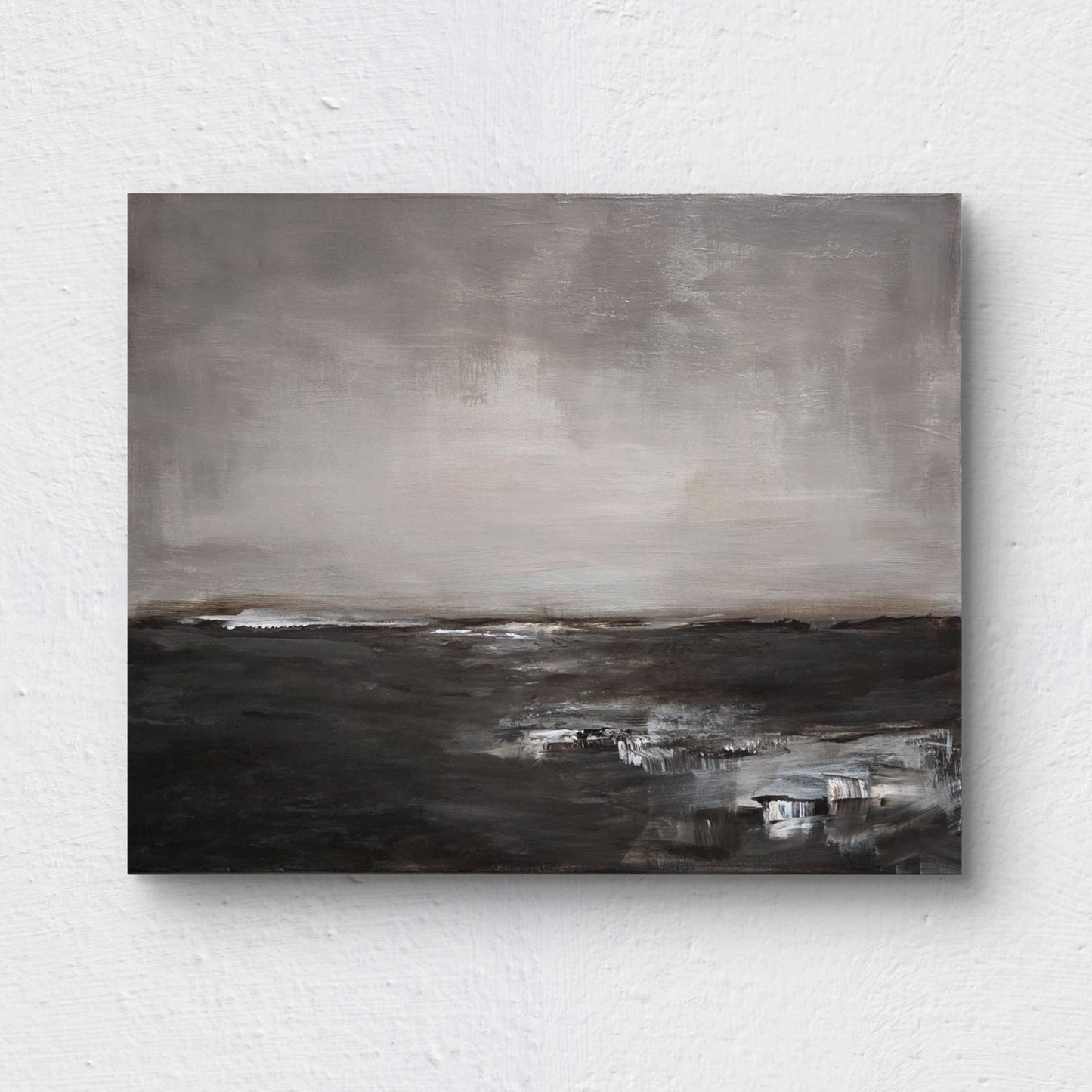 An abstract, minimalist landscape painting, rendered in muted shades of gray, white, and deep brown, suggesting a wide, bleak expanse of land or water meeting a vast, overcast sky. Painting is on a white wall.