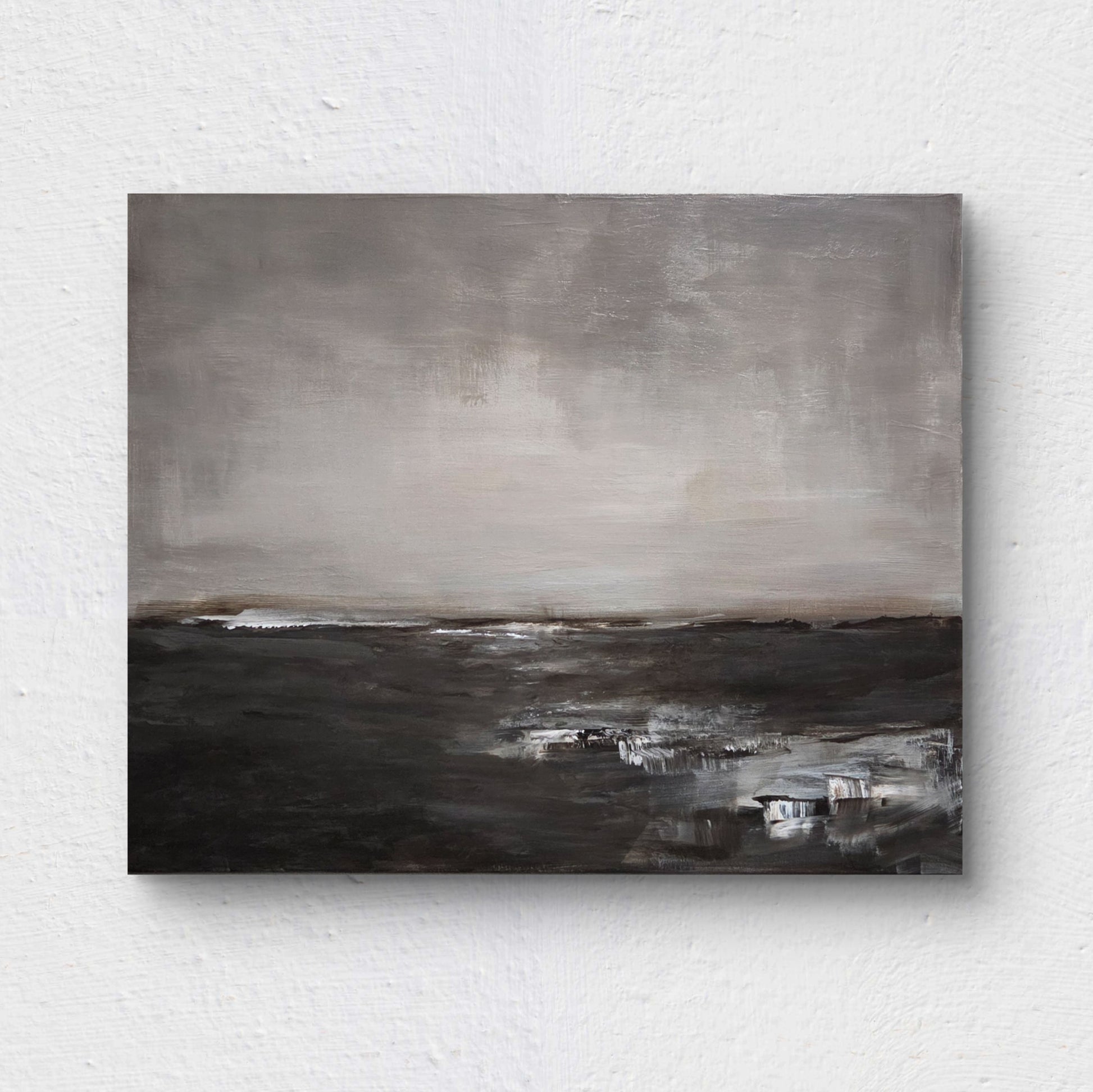An abstract, minimalist landscape painting, rendered in muted shades of gray, white, and deep brown, suggesting a wide, bleak expanse of land or water meeting a vast, overcast sky. Painting is on a white wall.
